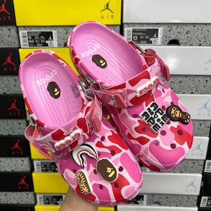 May include: A pair of pink Crocs clogs with a red and pink camouflage design. The clogs feature various decorative charms, including a shark, a brown ape head, and the text "APE NEW APE".