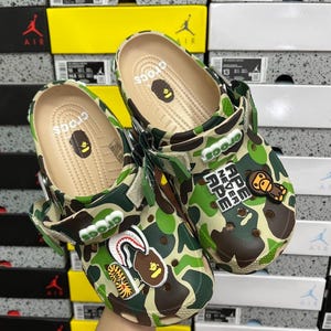 May include: Camouflage Crocs clogs with decorative charms. The clogs have a green, brown, and tan camouflage pattern. One clog features the text "APE NEVER APE" and a cartoon ape charm. The other clog has a shark and ape charm.
