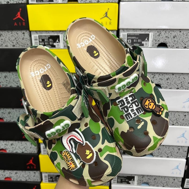 May include: Camouflage Crocs clogs with decorative charms. The clogs have a green, brown, and tan camouflage pattern. One clog features the text "APE NEVER APE" and a cartoon ape charm. The other clog has a shark and ape charm.