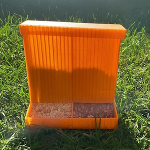 May include: An orange bird feeder with two compartments, filled with birdseed. The feeder is made of plastic and has a rectangular shape with vertical lines on the sides. It is sitting on green grass.