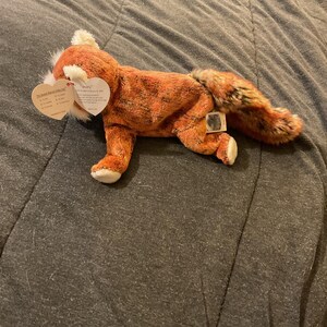 Könnte beinhalten: Ein Plüschfuchs mit orange-braunem Fell, weißen Akzenten und einem flauschigen Schwanz. Das Spielzeug hat ein herzförmiges Etikett. Der Fuchs liegt auf einer grauen Oberfläche.
