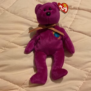 Könnte beinhalten: Ein lila Plüschteddybär mit schwarzer Nase und einem goldenen Band um den Hals. Der Bär hat ein kleines gesticktes Design auf der Brust und ein rotes Etikett an seinem Ohr.
