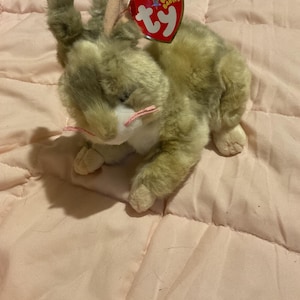 May include: A plush toy rabbit with a Ty heart-shaped tag. The rabbit has a light brown and tan fur, pink whiskers, and pink inner ears. The tag reads "Original Beanie Babies".