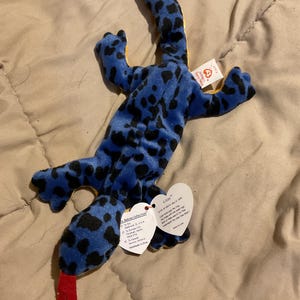 Puede incluir: Un juguete de peluche azul con forma de lagarto con manchas negras y una lengua roja. El juguete tiene una parte inferior amarilla y una etiqueta que dice "Lizzy". El juguete es parte de la colección Beanie Babies.