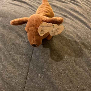 May include: A brown plush dachshund dog toy with a tag attached. The toy has a long body, short legs, and floppy ears. The tag reads "The Beanie Babies Collection".