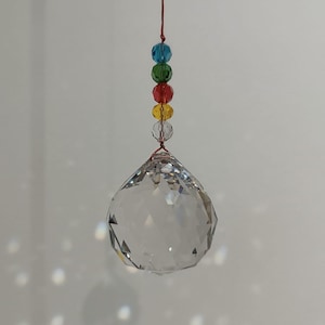Może przedstawiać: Przezroczysty kryształowy kulisty łapacz słońca z fasetowaną powierzchnią, zawieszony na czerwonym sznurku. Nad kulą kolorowe koraliki w kolorze niebieskim, zielonym, czerwonym, żółtym i przezroczystym. Tło to gładka biała ściana.