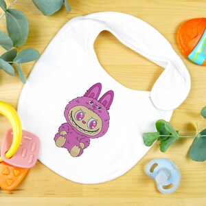 White baby bib with a pink kawaii monster embroidery design, styled with baby toys and eucalyptus, shown as a mockup for a digital embroidery file download