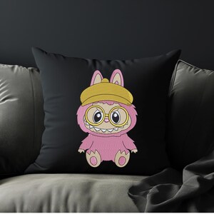 Black throw pillow on a sofa featuring a pink kawaii monster embroidery design with glasses and a yellow beanie – home decor mockup for a digital embroidery file