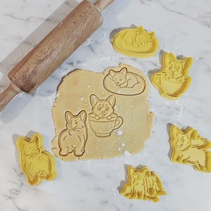 May include: A wooden rolling pin and several yellow plastic cookie cutters shaped like Corgi dogs in various poses, including sitting in a teacup and sleeping on a bed, pressed into a flat piece of light tan cookie dough on a white surface.
