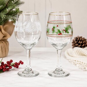 May include: Two clear glass wine glasses with gold rims. The left glass features a white etched holly leaf pattern. The right glass displays a painted design of green holly leaves and red berries, framed by two horizontal dotted red and green lines.