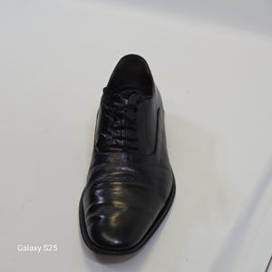 Puede incluir: Un zapato oxford de cuero negro con una puntera redondeada y lisa, un sistema de cordones cerrado con cordones negros finos y un tac&oacute;n bajo. El zapato tiene un acabado pulido y una silueta estructurada, mostrando un dise&ntilde;o cl&aacute;sico.