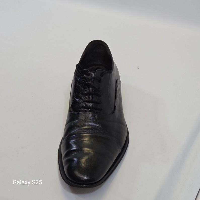 Puede incluir: Un zapato oxford de cuero negro con una puntera redondeada y lisa, un sistema de cordones cerrado con cordones negros finos y un tac&oacute;n bajo. El zapato tiene un acabado pulido y una silueta estructurada, mostrando un dise&ntilde;o cl&aacute;sico.