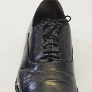 Puede incluir: Un zapato oxford de cuero negro con punta redondeada, sistema de cordones cerrados con cordones negros finos y un acabado liso y pulido. El zapato muestra pliegues naturales visibles en el empeine y una silueta estructurada.