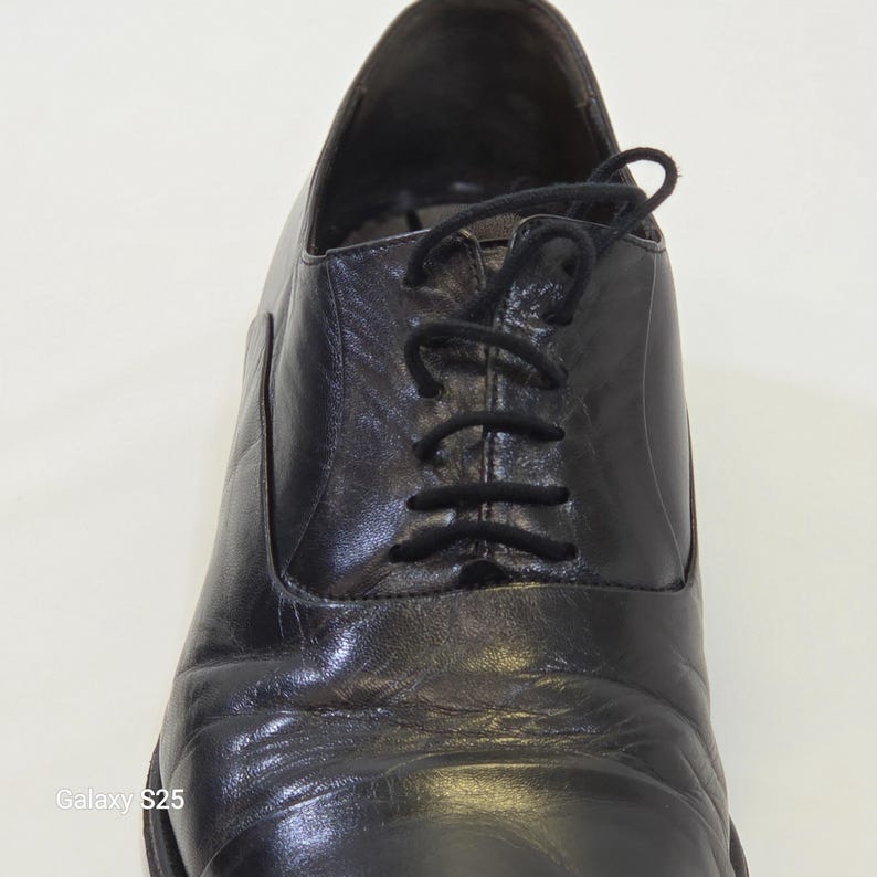 Puede incluir: Un zapato oxford de cuero negro con punta redondeada, sistema de cordones cerrados con cordones negros finos y un acabado liso y pulido. El zapato muestra pliegues naturales visibles en el empeine y una silueta estructurada.