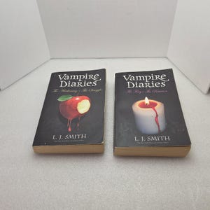Könnte beinhalten: Zwei Taschenbücher mit dem Titel "Vampire Diaries" von L. J. Smith. Ein Cover zeigt einen roten Apfel mit einem Biss, das andere eine Kerze mit tropfendem Wachs. Beide Bücher haben schwarze Einbände.