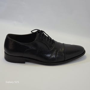 Puede incluir: Un zapato oxford de cuero negro con dise&ntilde;o de puntera, tac&oacute;n bajo y cordones negros finos. El zapato tiene un acabado liso y pulido con ligeras arrugas en el empeine y una silueta estructurada, presentado desde una vista de perfil lateral.