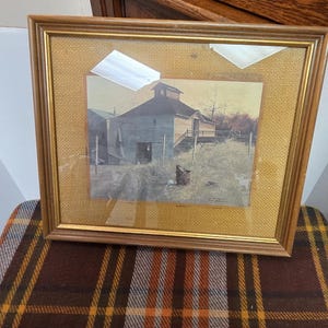 May include: A framed print of a rural scene featuring a weathered barn and surrounding landscape. The print is set within a gold-toned wooden frame and a textured, light-colored mat. The frame rests on a plaid patterned fabric.