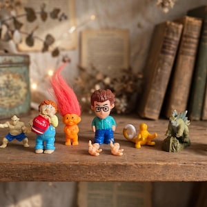 Puede incluir: Una colección de pequeñas figuras de plástico que incluye un humanoide pálido, una figura agachada, un personaje con un tarro de galletas rojo, un troll de pelo rosa brillante, una figura con gafas, dos bebés diminutos, una criatura amarilla y dos dinosaurios.