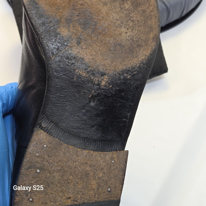 Puede incluir: La suela de un zapato de cuero negro que presenta una superficie desgastada y texturizada con signos visibles de uso. El tac&oacute;n est&aacute; construido con material apilado con peque&ntilde;os clavos met&aacute;licos visibles. El texto Galaxy S25 est&aacute; impreso en blanco.
