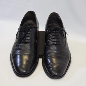 Puede incluir: Un par de zapatos oxford de cuero negro con un acabado liso y pulido, punteras redondeadas y cordones negros finos. Los zapatos tienen un sistema de cordones cerrado cl&aacute;sico y un tac&oacute;n bajo, presentados sobre un fondo claro y liso.