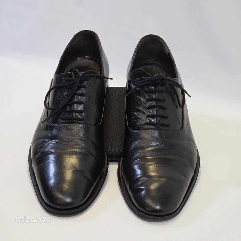 Puede incluir: Un par de zapatos oxford de cuero negro con un acabado liso y pulido, punteras redondeadas y cordones negros finos. Los zapatos tienen un sistema de cordones cerrado cl&aacute;sico y un tac&oacute;n bajo, presentados sobre un fondo claro y liso.