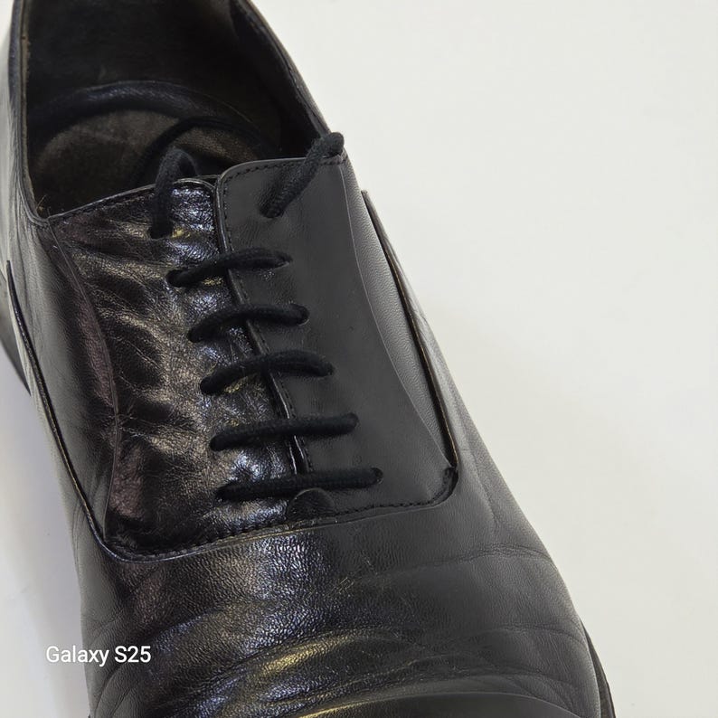 Puede incluir: Un zapato oxford de cuero negro con un sistema de cordones cerrado y cordones negros finos. El zapato tiene un acabado liso y pulido con pliegues naturales visibles en el empeine y una puntera estructurada. El texto Galaxy S25 es visible.