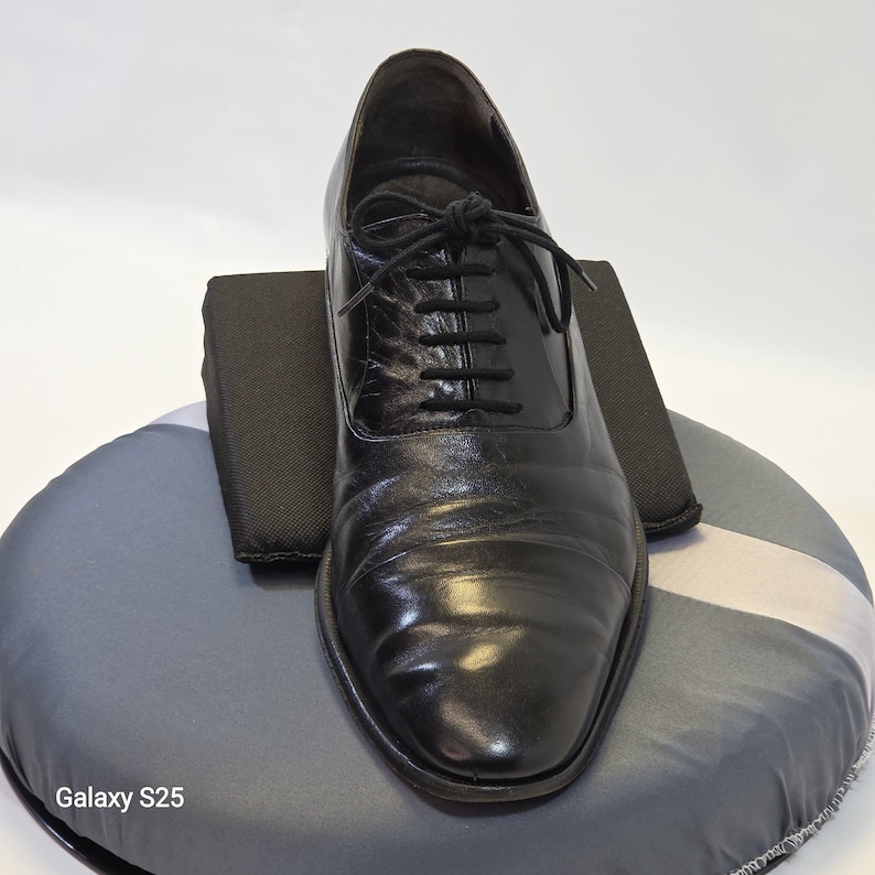 Puede incluir: Un zapato oxford de cuero negro con sistema de cordones cerrados y cordones negros finos. El zapato presenta un acabado liso y pulido con pliegues sutiles en el empeine y una punta redondeada. El texto Galaxy S25 es visible en la parte inferior izquierda.