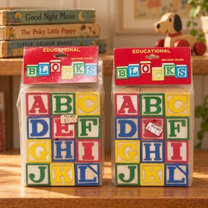 May include: Two packages of wooden alphabet blocks in clear plastic bags. Each package has a red header card with the text EDUCATIONAL BLOCKS and NON-TOXIC COLORS. The blocks feature letters A through L in red, green, blue, and yellow colors.