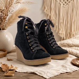 May include: A pair of black suede lace-up wedge boots featuring gold-toned metal eyelets, dark gray laces, and a textured tan rubber platform sole. The boots have a rounded toe and a high-top silhouette with a soft interior lining visible at the top.