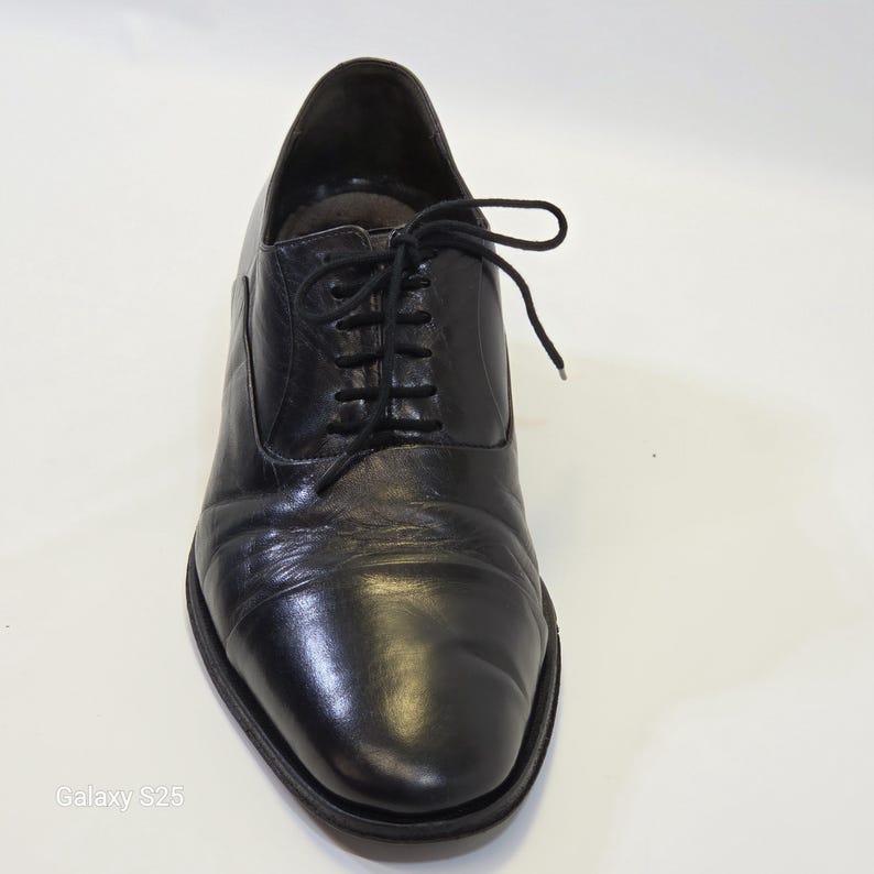 Puede incluir: Un zapato oxford de cuero negro con punta redondeada, acabado liso y cordones negros finos atados en un lazo. El zapato presenta una silueta cl&aacute;sica de perfil bajo con costuras visibles a lo largo de los paneles laterales y suela oscura.