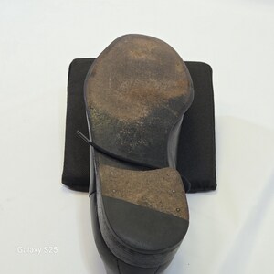 Puede incluir: Un zapato de cuero negro visto desde abajo, que revela una suela marr&oacute;n oscuro desgastada con marcas visibles y un tac&oacute;n apilado. El zapato presenta un cord&oacute;n negro que cruza la suela y est&aacute; colocado sobre un cuadrado de tela negra.