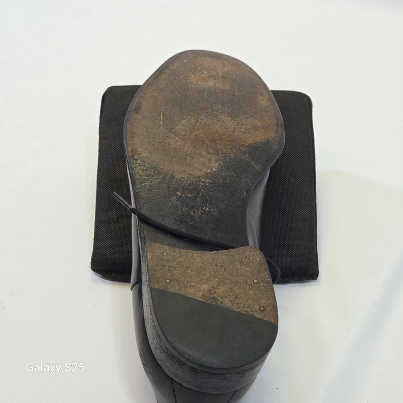 Puede incluir: Un zapato de cuero negro visto desde abajo, que revela una suela marr&oacute;n oscuro desgastada con marcas visibles y un tac&oacute;n apilado. El zapato presenta un cord&oacute;n negro que cruza la suela y est&aacute; colocado sobre un cuadrado de tela negra.