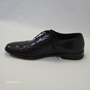 Puede incluir: Un zapato oxford de cuero negro con dise&ntilde;o de puntera, que presenta un cierre de cordones con cordones negros finos. El zapato tiene un acabado liso y pulido, un tac&oacute;n bajo y una silueta estructurada, presentado de perfil sobre una superficie lisa.