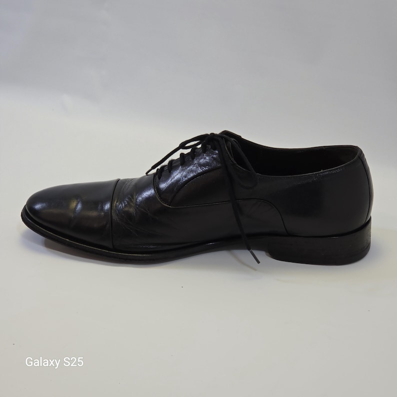 Puede incluir: Un zapato oxford de cuero negro con dise&ntilde;o de puntera, que presenta un cierre de cordones con cordones negros finos. El zapato tiene un acabado liso y pulido, un tac&oacute;n bajo y una silueta estructurada, presentado de perfil sobre una superficie lisa.