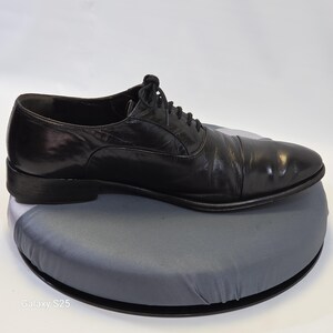 Puede incluir: Un zapato oxford de cuero negro con puntera, cordones negros finos y tac&oacute;n bajo. El zapato presenta un acabado liso y pulido con una silueta estructurada, mostrado de perfil sobre una base circular gris con el texto Galaxy S25.