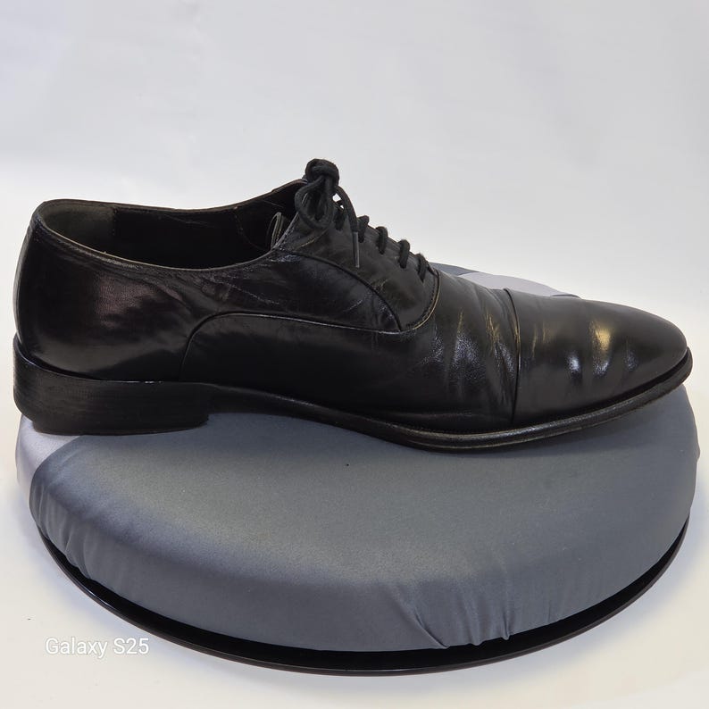 Puede incluir: Un zapato oxford de cuero negro con puntera, cordones negros finos y tac&oacute;n bajo. El zapato presenta un acabado liso y pulido con una silueta estructurada, mostrado de perfil sobre una base circular gris con el texto Galaxy S25.
