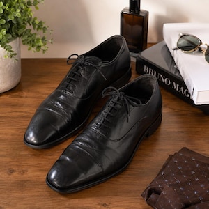 Puede incluir: Un par de zapatos oxford de cuero negro con dise&ntilde;o de puntera, cordones negros finos y tac&oacute;n bajo. Los zapatos tienen un acabado liso y pulido con costuras visibles a lo largo de los paneles de los ojales y una silueta estructurada.