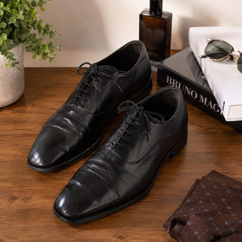 Puede incluir: Un par de zapatos oxford de cuero negro con dise&ntilde;o de puntera, cordones negros finos y tac&oacute;n bajo. Los zapatos tienen un acabado liso y pulido con costuras visibles a lo largo de los paneles de los ojales y una silueta estructurada.
