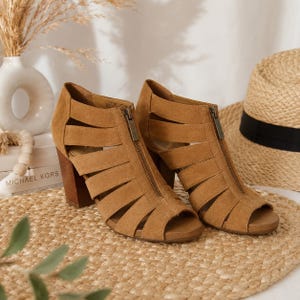 May include: A pair of tan suede peep-toe booties featuring a front zipper closure, multiple horizontal cut-out straps across the sides, and a stacked block heel. The footwear has a textured finish and a metallic zipper pull with a visible brand logo.