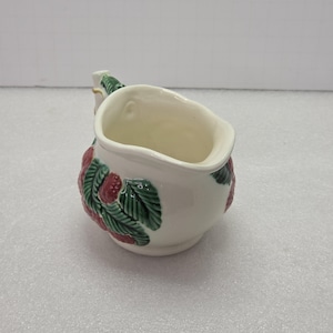 May include: A white ceramic creamer with a strawberry design. The creamer has a handle and a spout. The strawberries are red with green leaves. The creamer is approximately 4 inches tall.