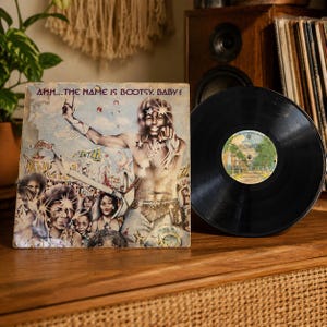 May include: A vinyl record album titled Ahh... The Name Is Bootsy, Baby! featuring a colorful illustrated cover of a musician with star-shaped glasses and a crowd, alongside a black vinyl record disc with a central label showing track listings.