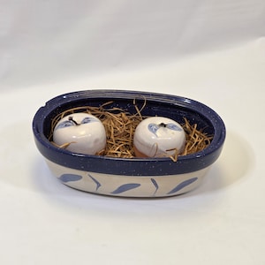 May include: A ceramic serving dish with a blue speckled rim and a cream-colored body. Inside, two white ceramic apples with blue leaf designs rest on a bed of straw. The dish has blue leaf-shaped accents.