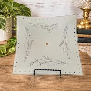 May include: A square, frosted glass light fixture shade featuring a central circular opening and a border of small, evenly spaced dots. The surface is decorated with four black line-art illustrations of wheat stalks arranged around the center.