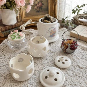 May include: A collection of white ceramic items including a lidded jar with a cat and flower basket illustration, a heart-cutout candle holder, a small bowl with scalloped edges, and two round lids with ventilation holes for fragrance.