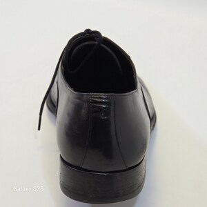 Puede incluir: Un zapato oxford de cuero negro con un acabado liso y pulido, un tac&oacute;n bajo y cordones negros finos. El zapato se muestra desde la parte posterior, destacando el contrafuerte redondeado, el detalle de la costura y el borde oscuro de la suela.