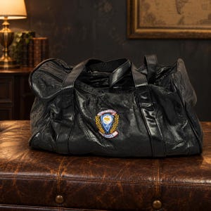 May include: A black leather duffel bag featuring a central embroidered patch with a blue shield, gold laurel leaves, and the text United States JCI Senate. The bag has two black handles and a zippered top closure with a textured, soft leather finish.