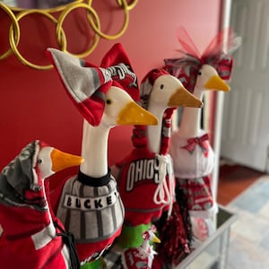 May include: Four decorative goose figurines dressed in red, white, and gray Ohio State themed outfits. The geese wear various fabric garments including hoodies, sweaters with the text BUCKEYES, and bows. Each goose has a white body and yellow beak.
