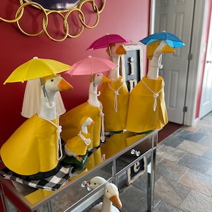 May include: A collection of decorative goose figurines, each adorned with a yellow raincoat, pearl necklace, and a colorful parasol. The geese are arranged on a mirrored table, creating a whimsical and charming display. A larger goose figurine stands on the floor.