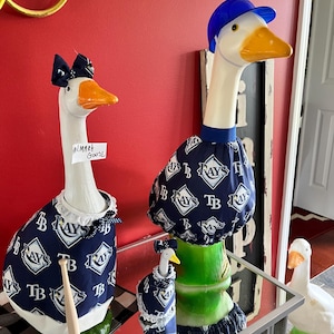 May include: Three white goose figurines wearing Tampa Bay Rays baseball team apparel. One goose wears a blue baseball cap. The geese are on a mirrored table with a red wall in the background. One goose has a small sign that says "Walmart Goose".