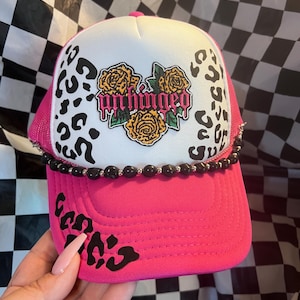 May include: A bright pink trucker hat featuring a white front panel with a black leopard print pattern. The center displays a graphic of three yellow roses with green leaves and the word unhinged in pink text. A black beaded chain accents the brim.